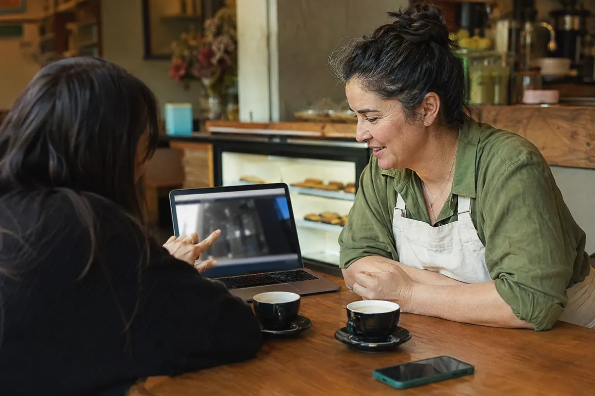 A person showing a short screen recording on a laptop to a small business owner across a cafe table, the owner leaning in interested, warm light