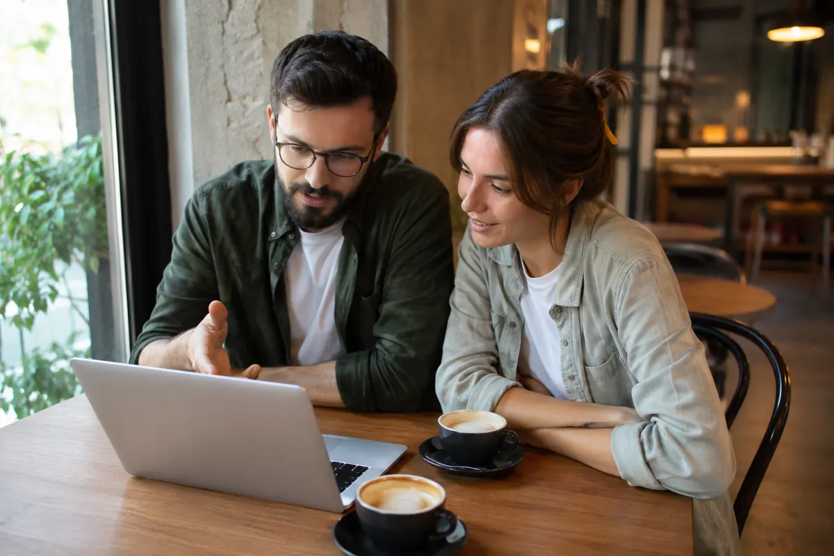 A freelancer and a small business owner sitting together at a cafe table looking at a laptop screen, discussing a workflow, warm natural light
