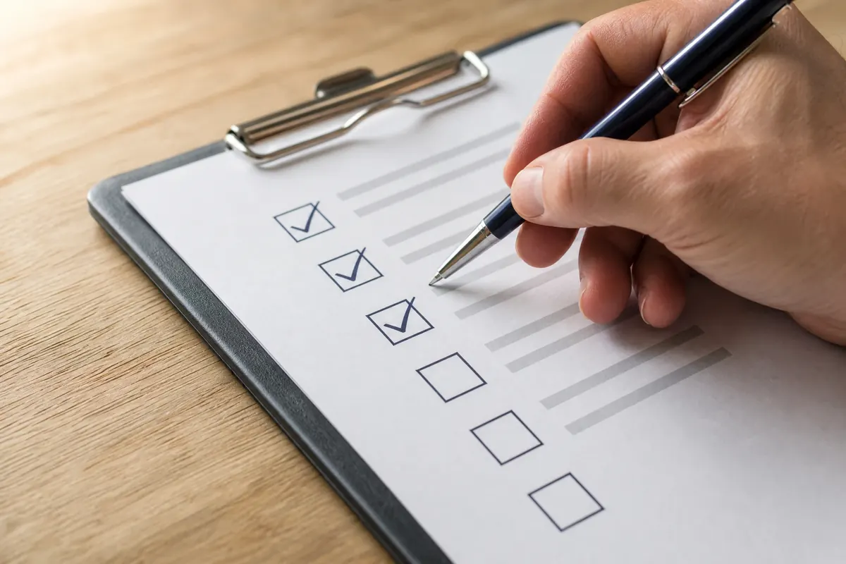 A hand holding a pen ticking boxes on a printed checklist attached to a clipboard, on a tidy desk in warm natural light